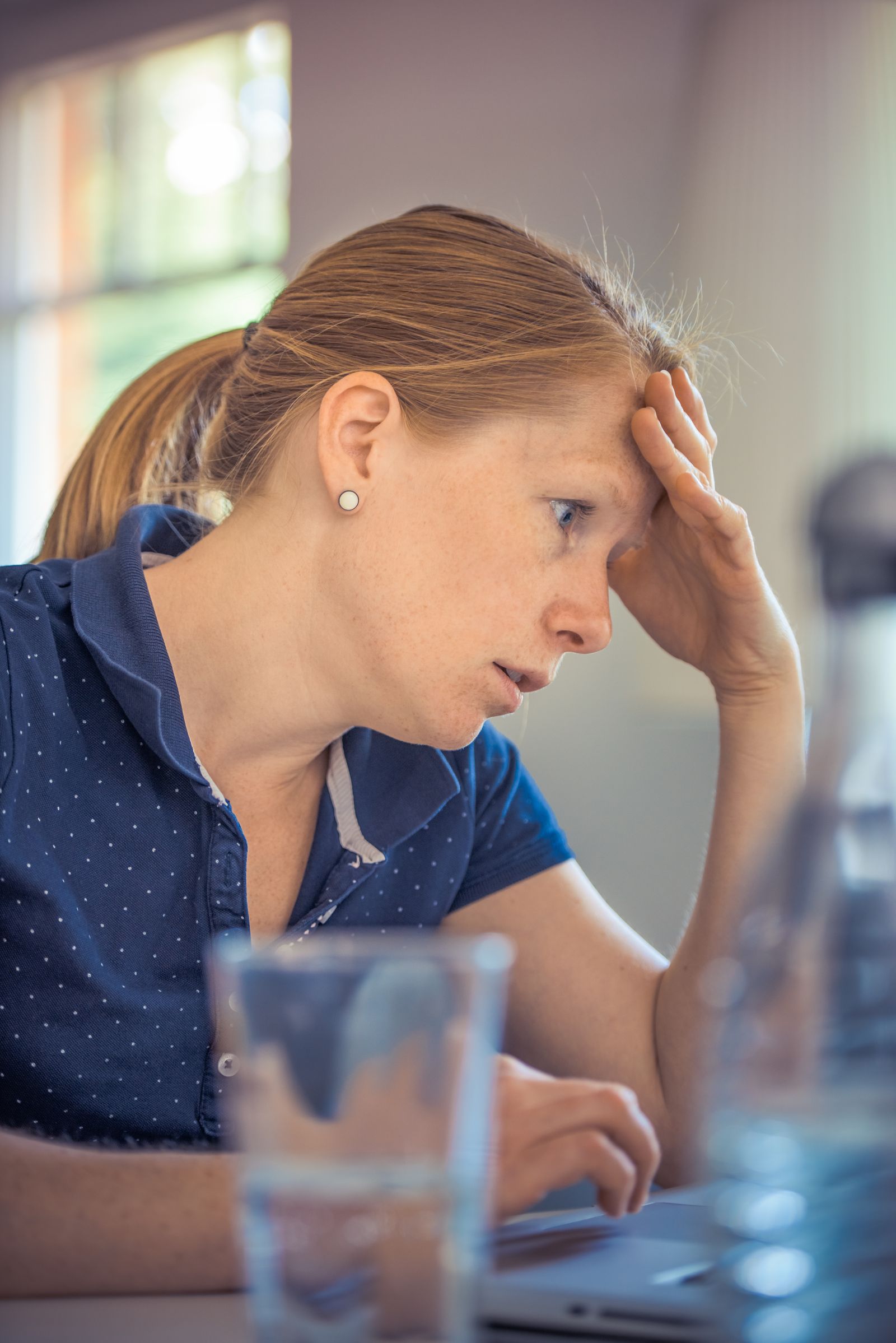 Eine Frau in gepunktetem Poloshirt stützt den Kopf in die Hand und blickt auf einen Laptop, im Vordergrund ein Glas.